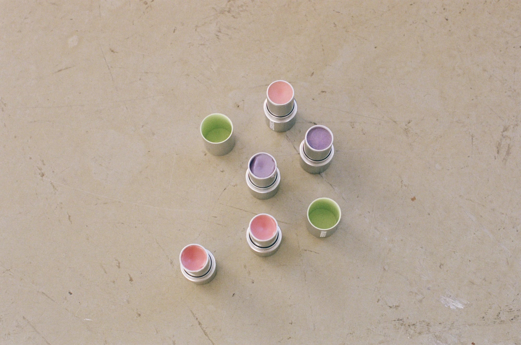 Six small cosmetic pots with their caps off, arranged on a textured beige surface. The pots contain pastel shades of pink, green, and purple.